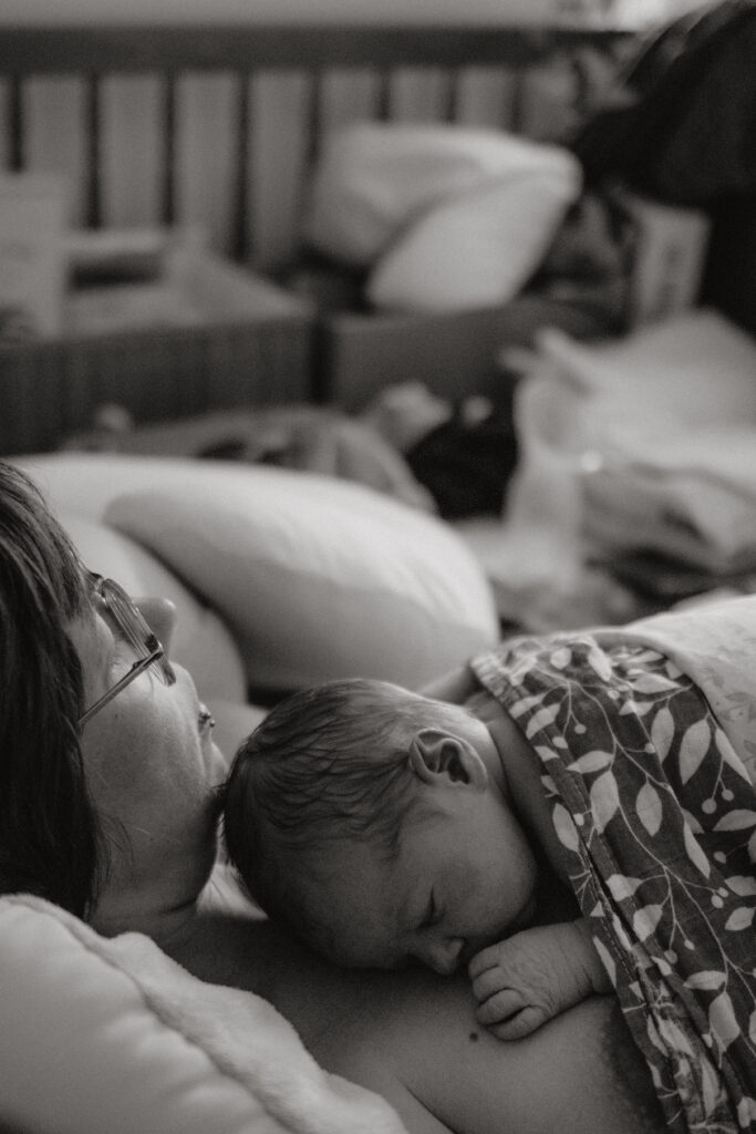 Eine Frau liegt mit einem neugeborenen Baby auf der Brust im Bett, im Hintergrund sind verschiedene Gegenstände zu sehen.